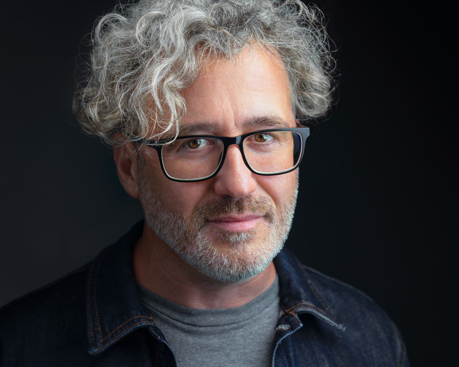 Business Headshot of a man with glasses, dark jean jacket and blue shirt on a dark background taken by Kalin Tabov Photography in Irvine, CA.